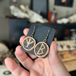 May include: Round wooden earrings with bronze-colored hooks. Each earring features a dark, textured background with a carved rune symbol. The earrings are displayed on a black card, held by a hand.