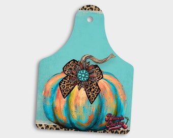 May include: A turquoise cutting board with a painted pumpkin design. The pumpkin has a brown stem, a turquoise and orange body, and a leopard print bow with a turquoise jewel. The text "Black Cherry" is printed on the board.