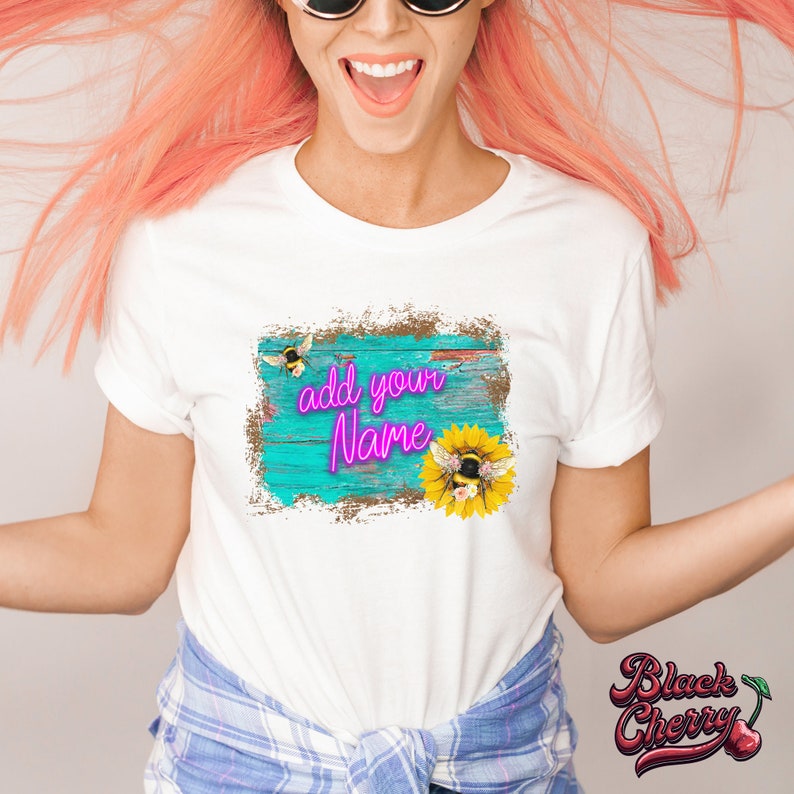 May include: White t-shirt with a distressed turquoise blue background with a wooden plank texture. The text "add your name" is in pink neon lettering. A sunflower with a bee on it and a bee flying above the sunflower are on the right side of the design.
