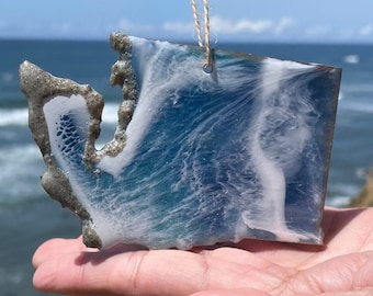 May include: A blue and white resin ornament shaped like a wave, hanging from a twine string. The ornament is being held in a hand against a blurred background of a beach and ocean.