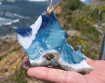 Puede incluir: Un adorno de ola de océano de resina azul y blanca con una cuerda atada. El adorno tiene forma de ola y tiene una cresta blanca y un valle azul. El adorno está siendo sostenido por una mano contra un fondo de playa y océano.
