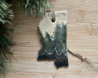 May include: A ceramic ornament shaped like the state of Mississippi with a blue and white glaze depicting a forest scene.