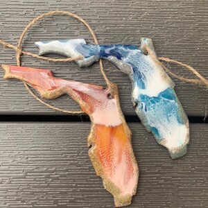May include: Two resin ornaments shaped like the state of Florida. One is blue, white, and gold, and the other is orange, pink, and gold.
