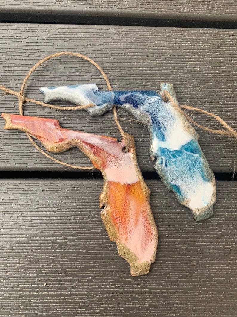 May include: Two resin ornaments shaped like the state of Florida. One is blue, white, and gold, and the other is orange, pink, and gold.