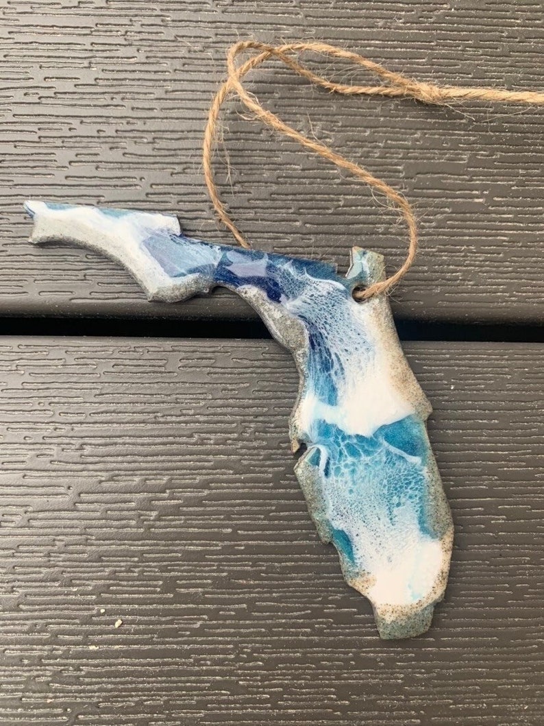 May include: A blue and white resin ornament shaped like the state of Florida. The ornament has a rustic twine hanger.