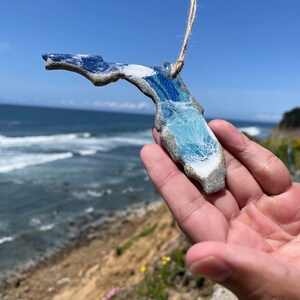 May include: A blue and white ceramic ornament shaped like the state of Florida. The ornament is hanging from a string and is being held in a hand against a backdrop of a beach and ocean.