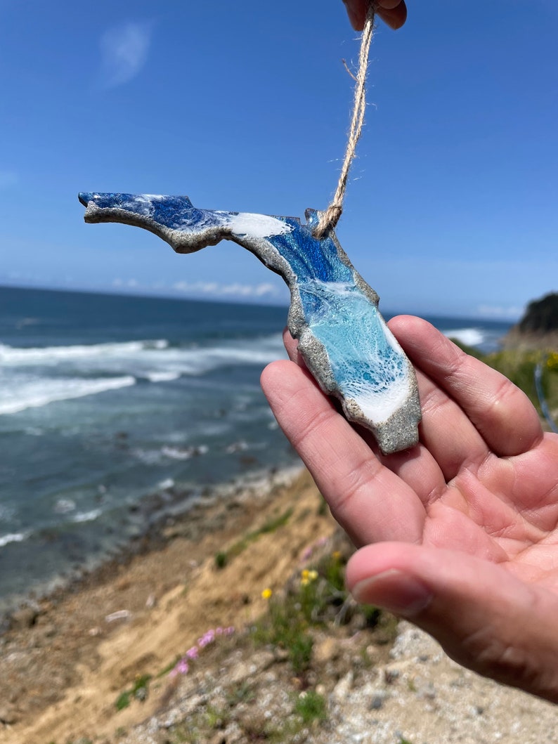 May include: A blue and white ceramic ornament shaped like the state of Florida. The ornament is hanging from a string and is being held in a hand against a backdrop of a beach and ocean.