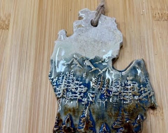 Puede incluir: Un adorno de cerámica con la forma del estado de Michigan, con un esmalte brillante que muestra un paisaje de montañas azules y pinos de color marrón oscuro. La parte superior es de color crema y tiene un cordel en el orificio.