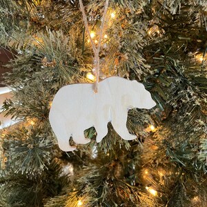 Puede incluir: Un adorno de oso de madera blanco colgado de un &aacute;rbol de Navidad con luces blancas.