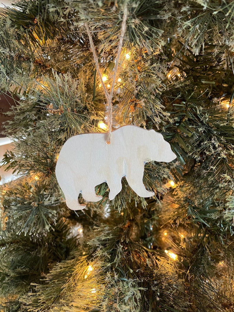Puede incluir: Un adorno de oso de madera blanco colgado de un &aacute;rbol de Navidad con luces blancas.