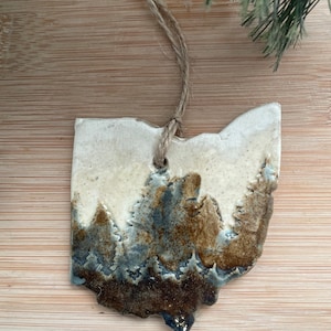 Puede incluir: Un adorno de cer&aacute;mica con forma del estado de Ohio con un esmalte marr&oacute;n y azul que representa una escena de bosque. El adorno est&aacute; colgado de una cuerda de yute.
