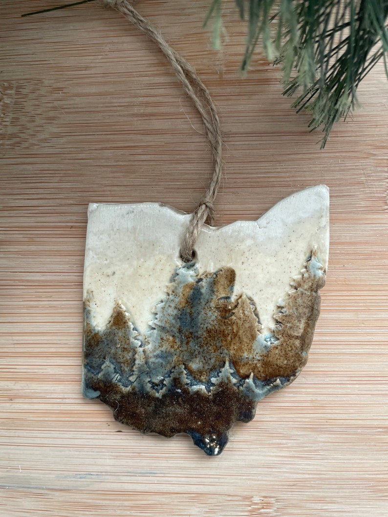 Puede incluir: Un adorno de cer&aacute;mica con forma del estado de Ohio con un esmalte marr&oacute;n y azul que representa una escena de bosque. El adorno est&aacute; colgado de una cuerda de yute.