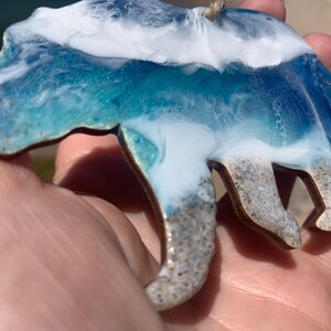 Puede incluir: Un contorno de oso de madera relleno de resina azul y blanca, que se asemeja a las olas del oc&eacute;ano. La resina tiene una textura brillante y el oso cuelga de una cuerda.