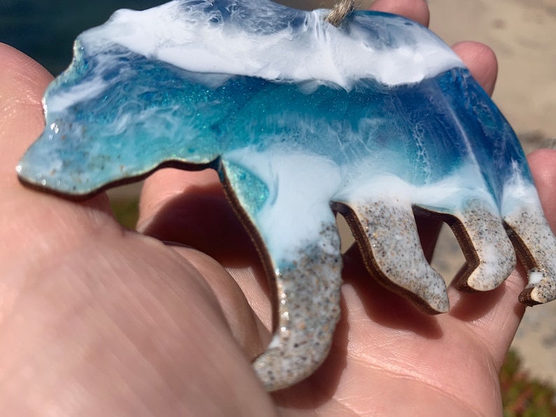 Puede incluir: Un contorno de oso de madera relleno de resina azul y blanca, que se asemeja a las olas del oc&eacute;ano. La resina tiene una textura brillante y el oso cuelga de una cuerda.