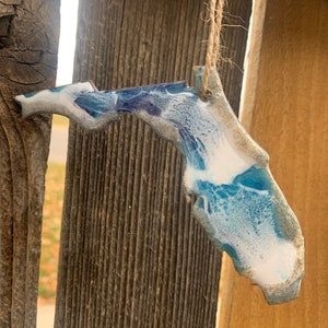 May include: A blue and white resin ornament shaped like the state of Florida. The ornament is hanging from a string and has a rustic wood background.