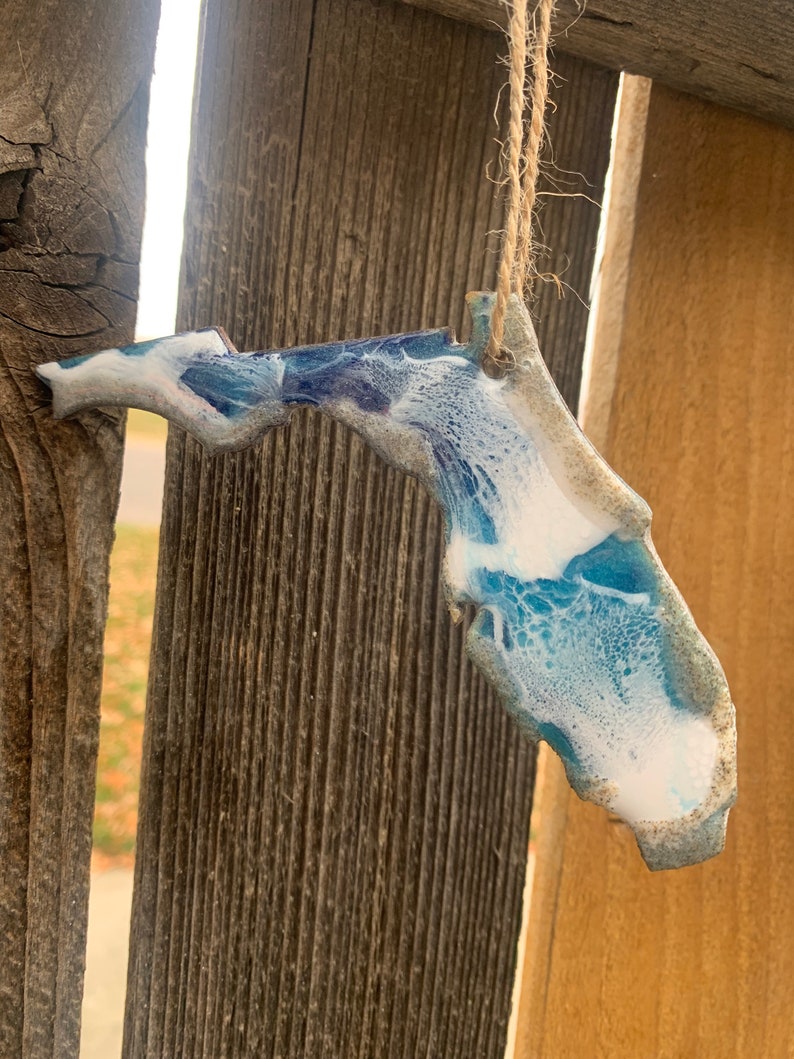 May include: A blue and white resin ornament shaped like the state of Florida. The ornament is hanging from a string and has a rustic wood background.