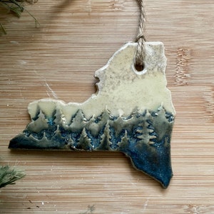 May include: A ceramic ornament shaped like a state with a blue and white forest scene. The ornament has a hole at the top for hanging.