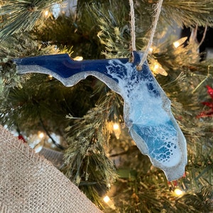 May include: A blue and white resin ornament shaped like the state of Florida. The ornament is hanging on a Christmas tree with a brown burlap ribbon.