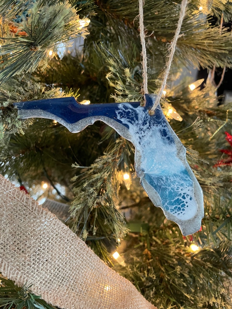 May include: A blue and white resin ornament shaped like the state of Florida. The ornament is hanging on a Christmas tree with a brown burlap ribbon.