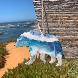 Puede incluir: Un adorno de oso de resina con un dise&ntilde;o de ola de oc&eacute;ano azul y blanco. El oso est&aacute; colgado de una cuerda en una superficie de madera.