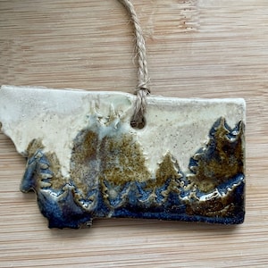 May include: A ceramic ornament shaped like the state of Montana with a white background and brown and blue glaze depicting a mountain range.