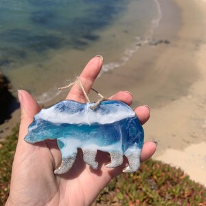 Puede incluir: Un adorno de oso de resina con un dise&ntilde;o de ola de oc&eacute;ano azul, blanco y verde. El oso est&aacute; de pie en una playa de arena.