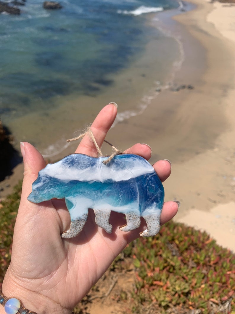 Puede incluir: Un adorno de oso de resina con un dise&ntilde;o de ola de oc&eacute;ano azul, blanco y verde. El oso est&aacute; de pie en una playa de arena.