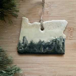 May include: A ceramic ornament shaped like the state of Oregon with a white background and a dark green forest scene at the bottom.