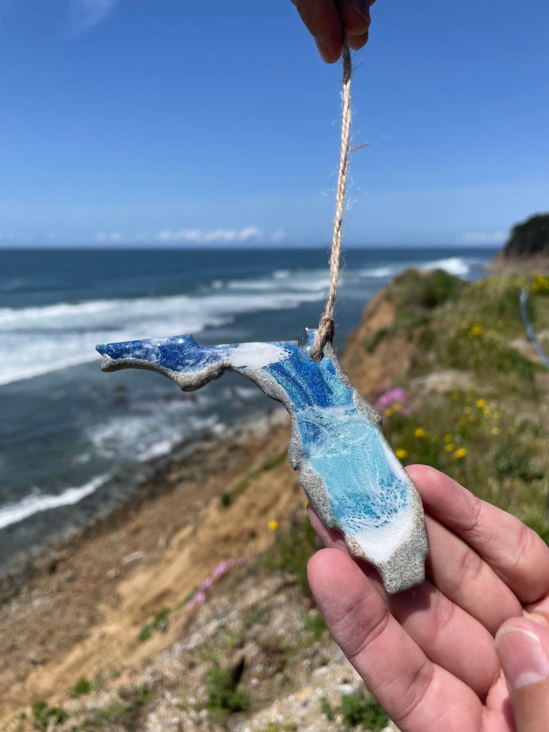 May include: A blue and white ceramic ornament shaped like the state of Florida, hanging from a twine string. The ornament is being held by a hand against a backdrop of a blue sky and ocean.