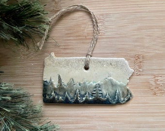 May include: A ceramic ornament shaped like the state of Pennsylvania with a blue and white glaze depicting a forest scene. The ornament has a twine hanger.