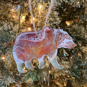 Puede incluir: Un adorno de oso de resina con un dise&ntilde;o marmoleado blanco y naranja. El adorno est&aacute; colgado de un &aacute;rbol de Navidad.
