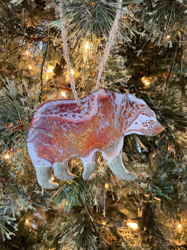 Puede incluir: Un adorno de oso de resina con un dise&ntilde;o marmoleado blanco y naranja. El adorno est&aacute; colgado de un &aacute;rbol de Navidad.