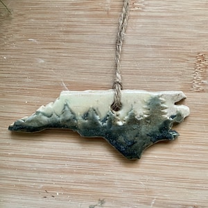 Puede incluir: Adorno de cerámica con la forma de Carolina del Norte, con una escena de bosque en tonos grises y verdes. Cuelga de una cuerda de hilo natural. Ideal para decoraciones festivas o como regalo único, mide unos 10 cm de largo.