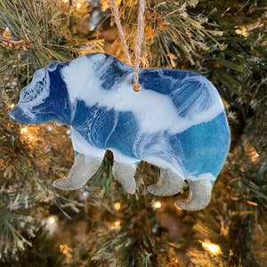 Puede incluir: Un adorno de oso de resina azul y blanco con detalles de purpurina. El oso est&aacute; colgado de un &aacute;rbol de Navidad.