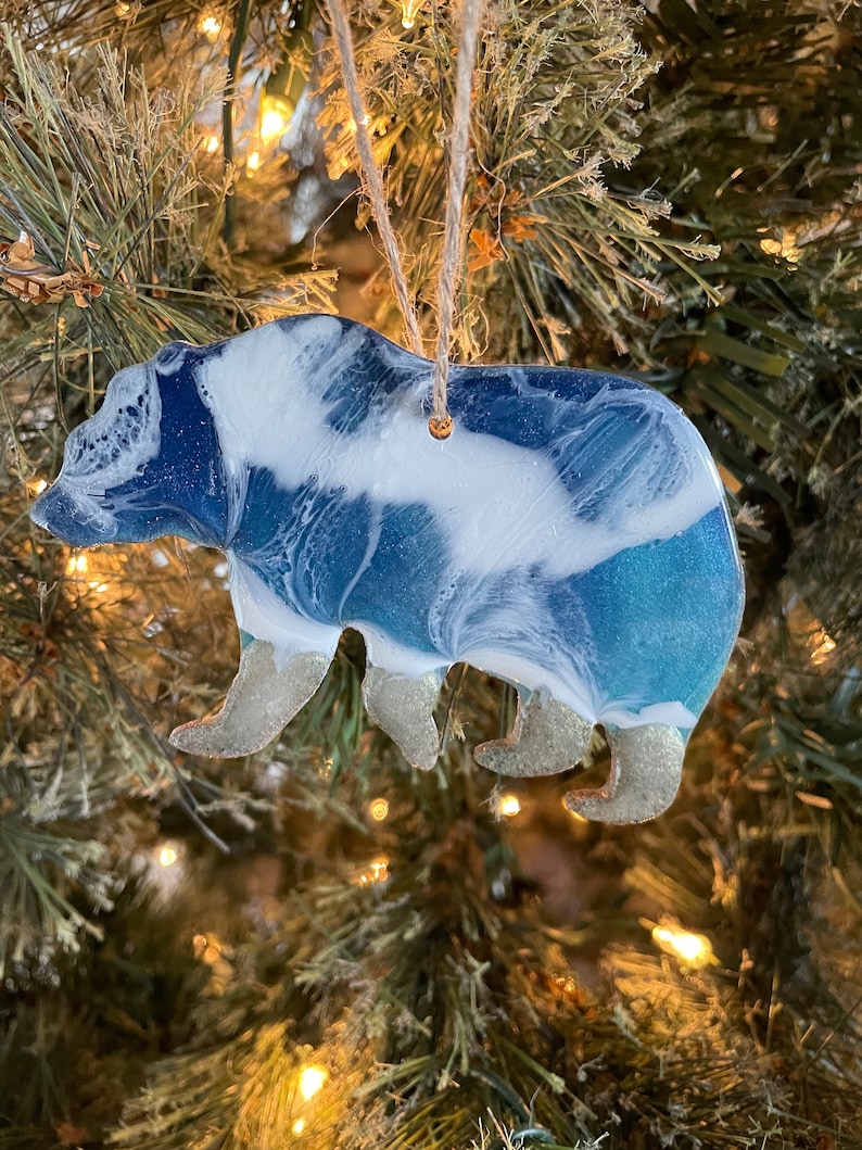 Puede incluir: Un adorno de oso de resina azul y blanco con detalles de purpurina. El oso est&aacute; colgado de un &aacute;rbol de Navidad.