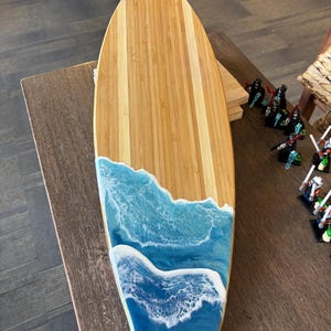 Könnte beinhalten: Ein holzförmiges Servierbrett in Surfbrettform. Die Oberseite besteht aus hellbraunem Bambus, während die untere Hälfte ein blau-weißes Harzdesign mit Meereswellen aufweist. Das Brett ist zum Servieren von Speisen konzipiert.