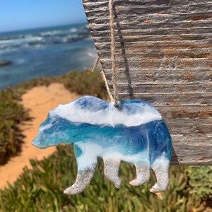 Puede incluir: Un adorno de oso de resina con un dise&ntilde;o de ola de oc&eacute;ano azul y blanco. El oso est&aacute; de pie en una playa de arena.