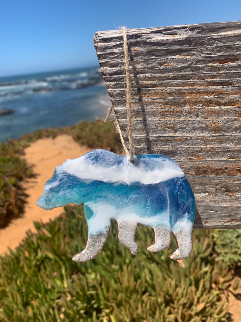 Puede incluir: Un adorno de oso de resina con un dise&ntilde;o de ola de oc&eacute;ano azul y blanco. El oso est&aacute; de pie en una playa de arena.