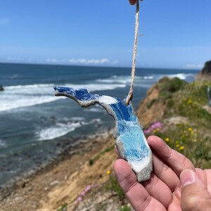 May include: A blue and white ceramic ornament shaped like the state of Florida, hanging from a twine string. The ornament is being held by a hand against a backdrop of a sandy cliff and ocean waves.