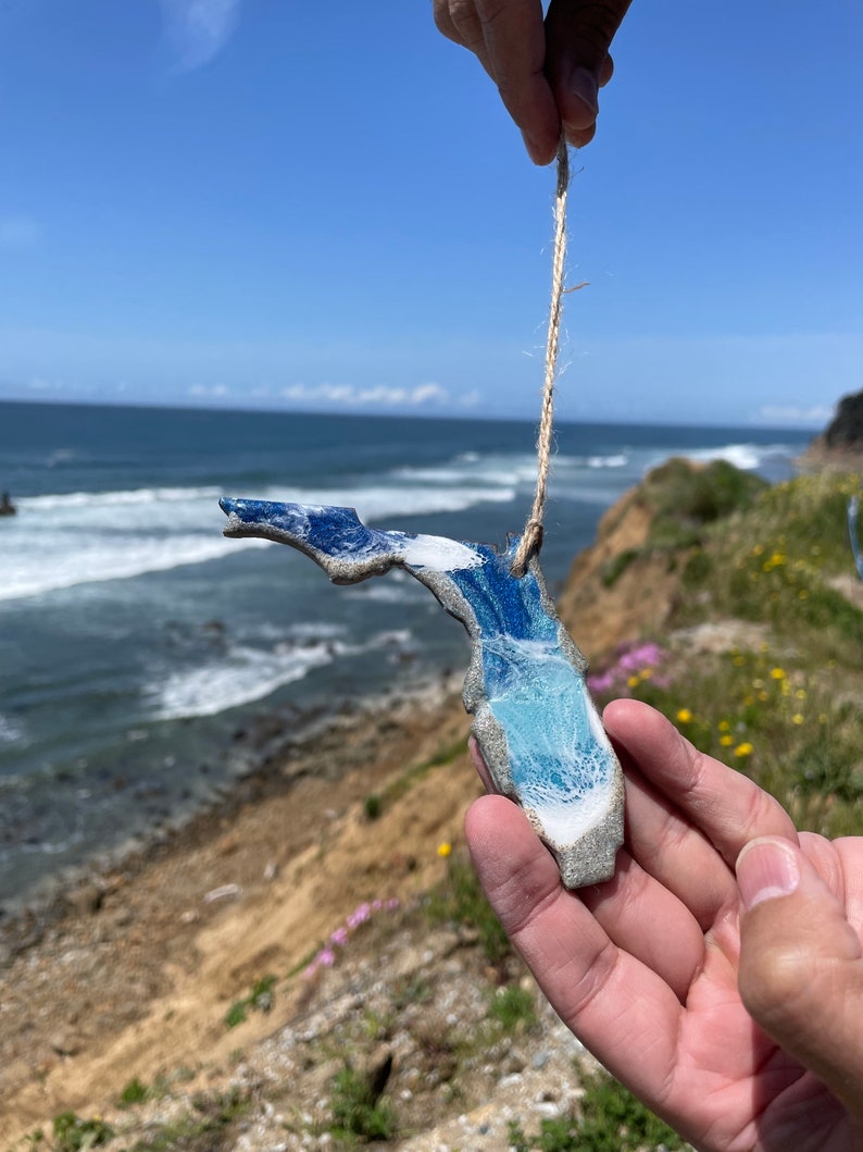 May include: A blue and white ceramic ornament shaped like the state of Florida, hanging from a twine string. The ornament is being held by a hand against a backdrop of a sandy cliff and ocean waves.