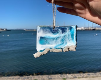 May include: A blue and white resin ornament with a rough, textured edge, resembling a wave. It is hanging from a twine string.