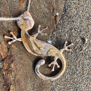 May include: A gold and white marbled gecko pendant with a rope necklace. The gecko is curled up with its tail wrapped around its body.