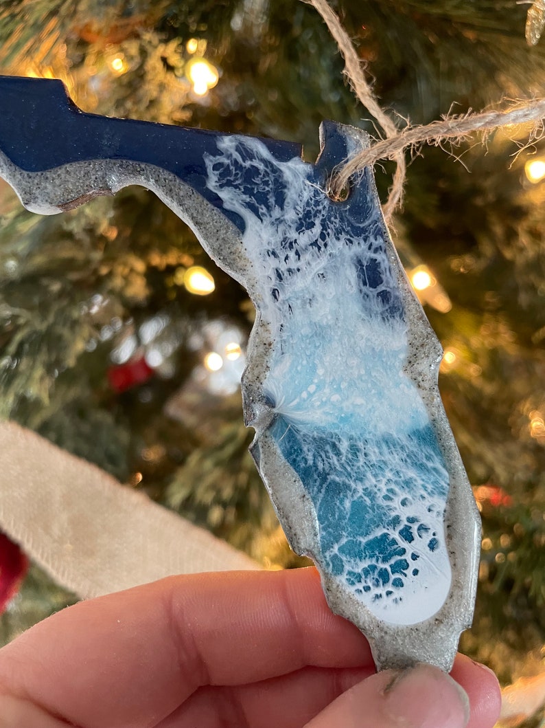 May include: A blue and white resin ornament shaped like the state of Florida. The ornament has a white wave design and a glittery finish.