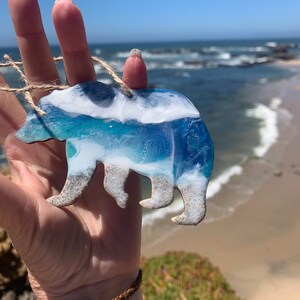 Puede incluir: Un adorno de oso de resina azul y blanco con un dise&ntilde;o de ola. El oso sostiene un peque&ntilde;o trozo de madera a la deriva en su boca.