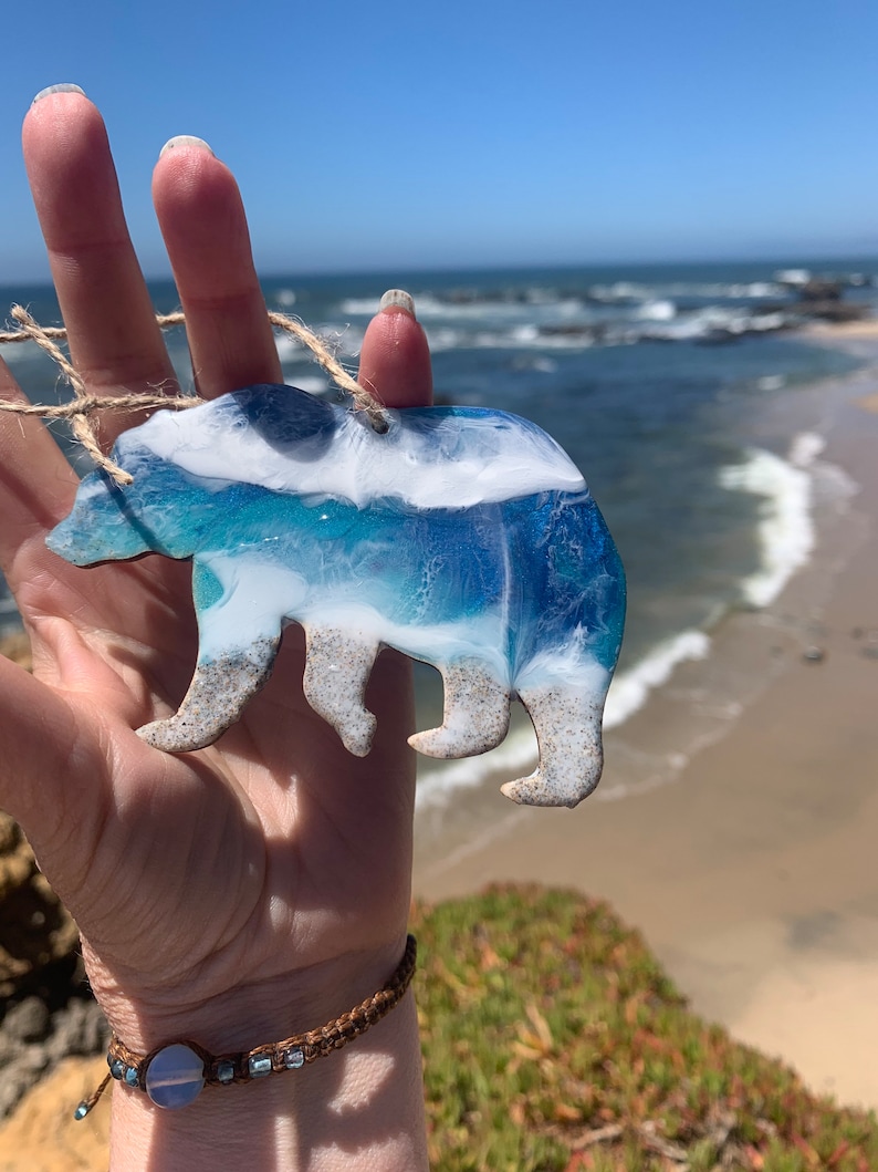Puede incluir: Un adorno de oso de resina azul y blanco con un dise&ntilde;o de ola. El oso sostiene un peque&ntilde;o trozo de madera a la deriva en su boca.