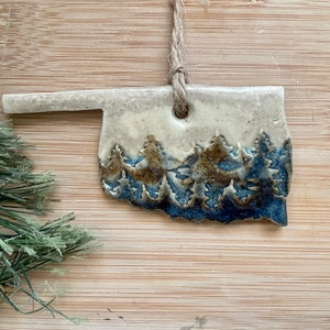 May include: A ceramic ornament shaped like the state of Oklahoma with a blue and brown glaze depicting a forest scene.
