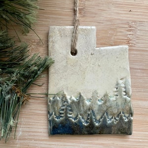 May include: A white ceramic ornament shaped like the state of Utah with a blue and green silhouette of a forest on the bottom.