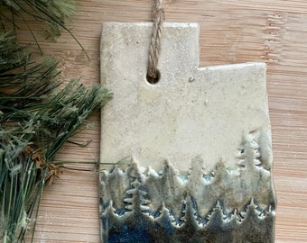 May include: A white ceramic ornament shaped like the state of Utah with a blue and green silhouette of a forest on the bottom.