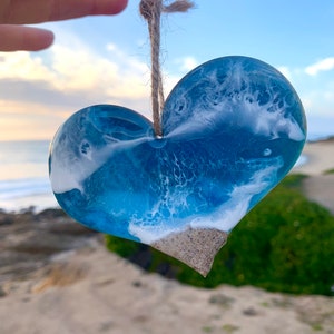 Può includere: Ornamento a forma di cuore in resina con un design a tema spiaggia. L'ornamento presenta sfumature di blu e bianco, che imitano le onde dell'oceano, e un fondo sabbioso. Un anello di spago naturale è attaccato per appenderlo.