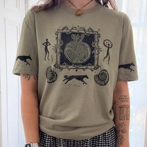 May include: A sage green t-shirt featuring a black ink-style graphic print. The central design shows a pomegranate inside a square frame, surrounded by stylised figures, running dogs, and spiral shells. The print extends onto both short sleeves.